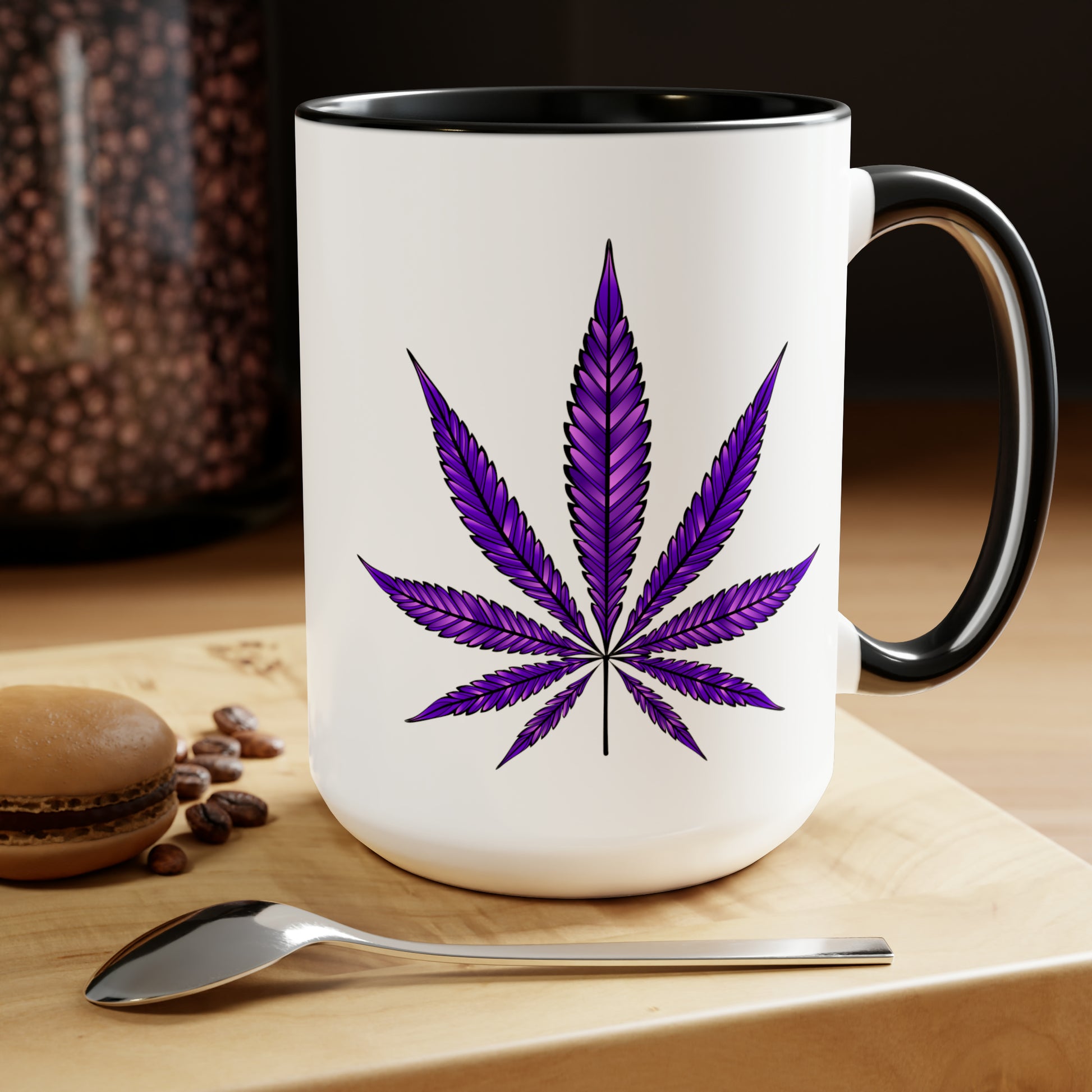 A Purple Haze Marijuana Coffee Mug with a leaf design on it, placed on a wooden table next to a spoon and a macaron.