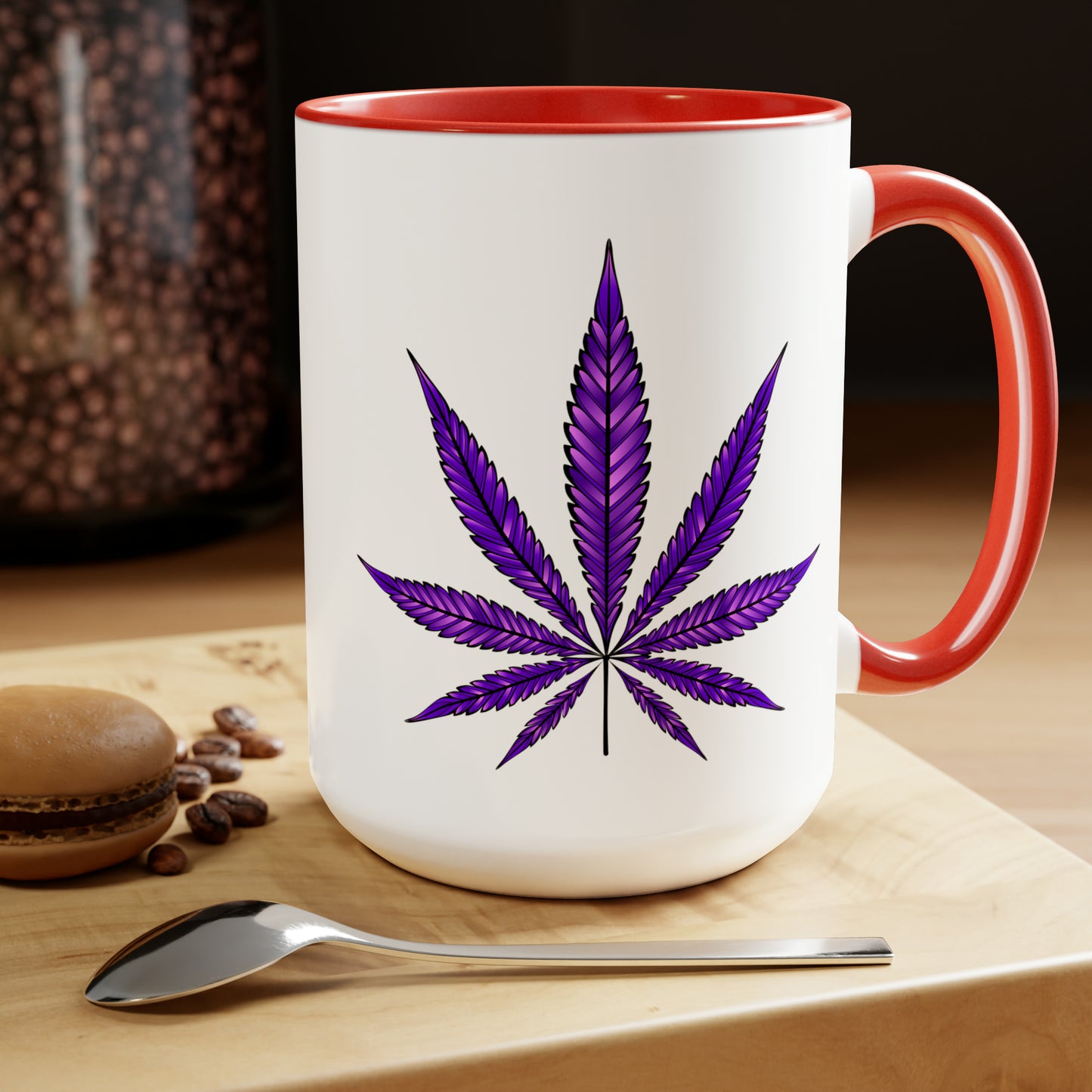 The Purple Haze Marijuana Coffee Mug with cannabis leaf design on wooden table, accompanied by coffee beans, a spoon, and a macaron.