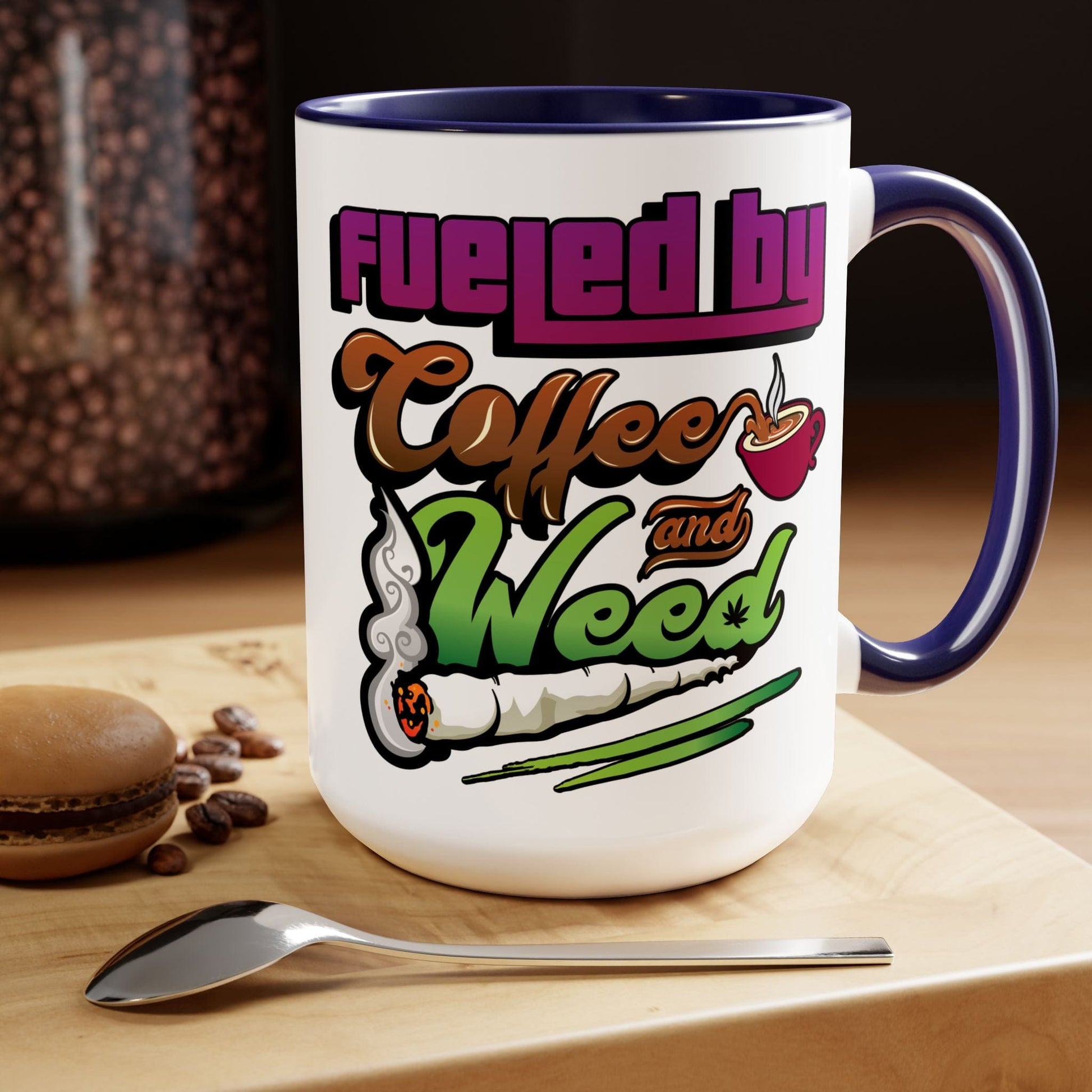 The Fueled by Coffee and Weed 15oz Coffee Mug is a white ceramic mug featuring the phrase "Fueled by Coffee and Weed" with a coffee cup and joint graphic, photographed on a wooden surface with a macaron, coffee beans, and spoon.