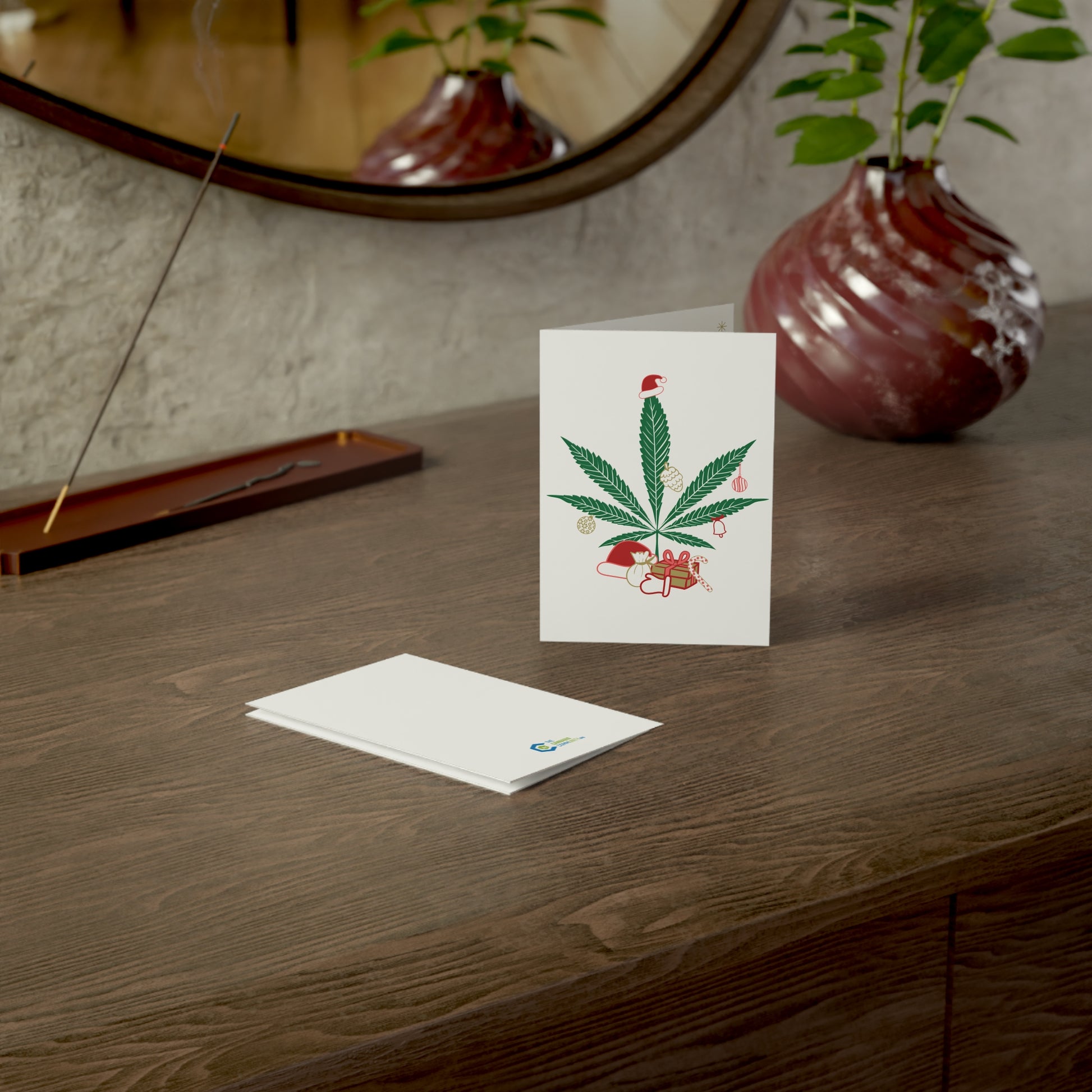 A Mary Kushmas + Dope New Year Marijuana Tree Christmas Greeting Card, featuring a floral design, printed on FSC certified paper, stands on a wooden table, accompanied by an envelope and decorative items including a vase with plants and a round mirror.