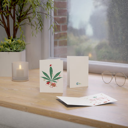 A Mary Kushmas + Dope New Year Marijuana Tree Christmas Greeting Card with a floral design, printed on FSC certified paper, stands on a wooden table beside candles, glasses, and another card near a foggy window.