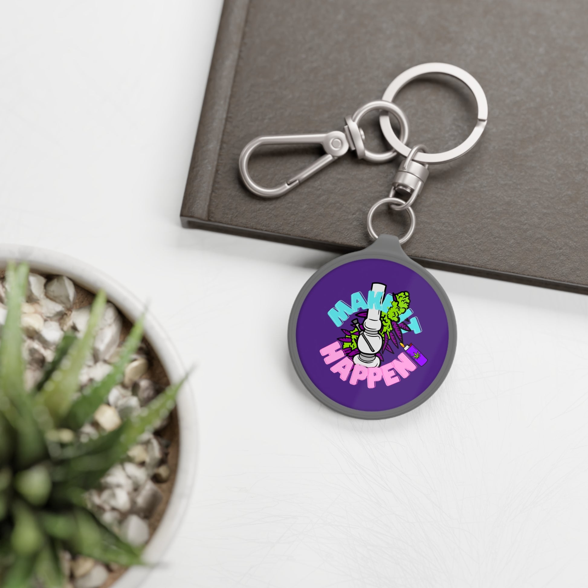 A keychain with a "Make It Happen Cannabis Keyring Tag" design next to a small potted plant and a closed notebook on a white surface.