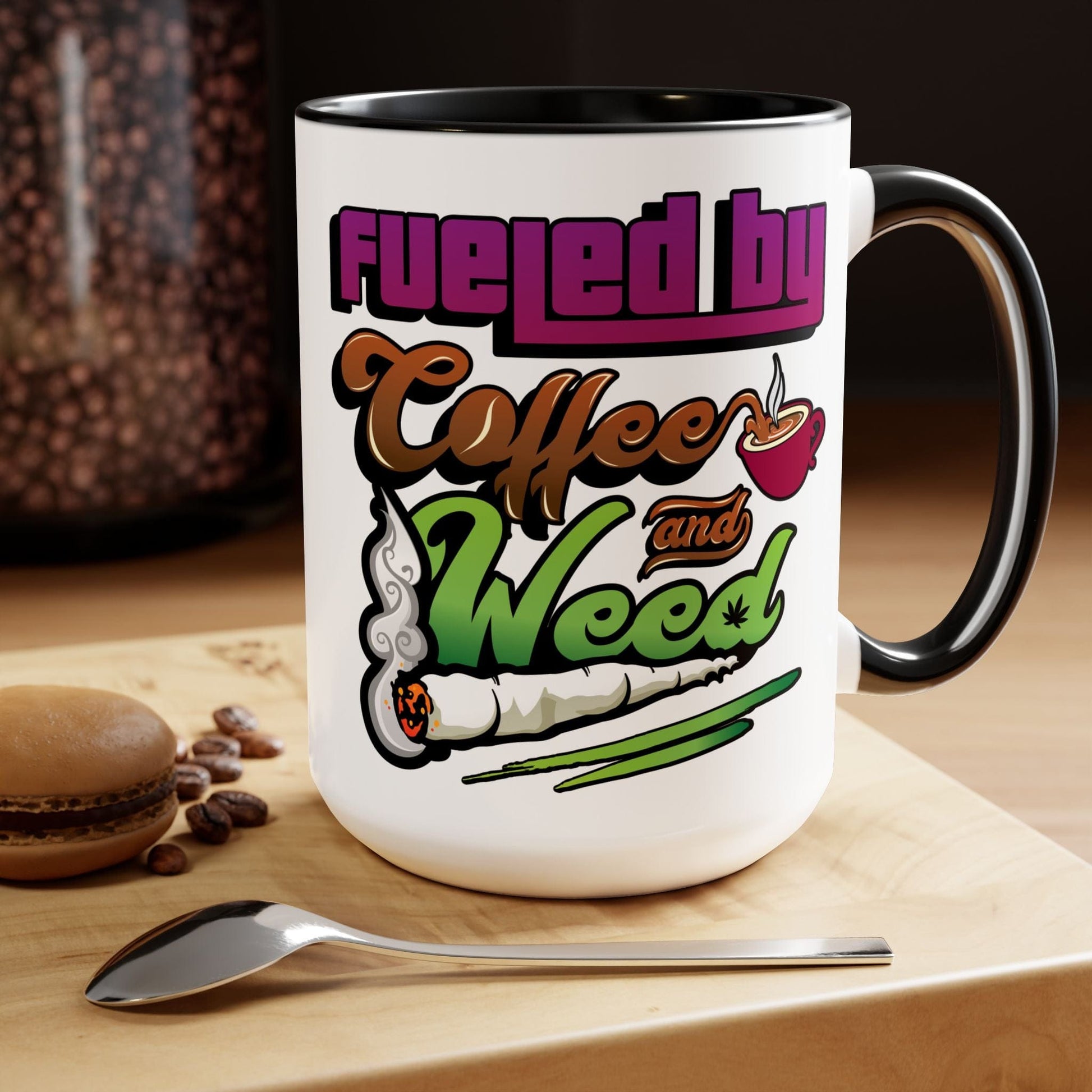 The Fueled by Coffee and Weed 15oz Coffee Mug, featuring a coffee cup and rolled joint graphic with the phrase "Fueled by Coffee and Weed," sits on a wooden surface beside a spoon and macaron.