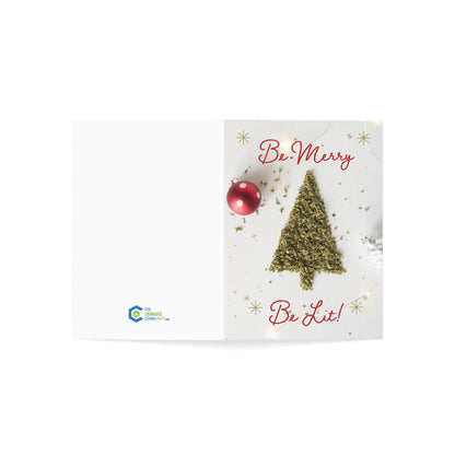 Open greeting card with a Christmas tree made of cannabis, the phrase 'Be Merry Be Lit' in red, and a red ornament, plus the logo of The Cannabis Community on the blank side.