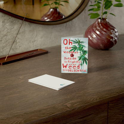 A Weed is So Delightful greeting card on a wooden table with text reading "oh the weather outside is frightful but the weed is so delightful," next to a red vase and a plant is made from sustainable paper.