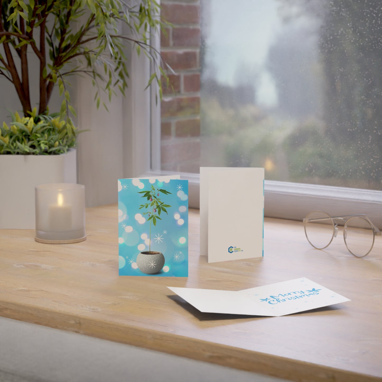 A serene workspace with a Potted Christmas Marijuana Plant Merry Christmas Greeting Card, blank envelope, glasses, and a lit candle on a wooden table by a window with a view of a rainy day.