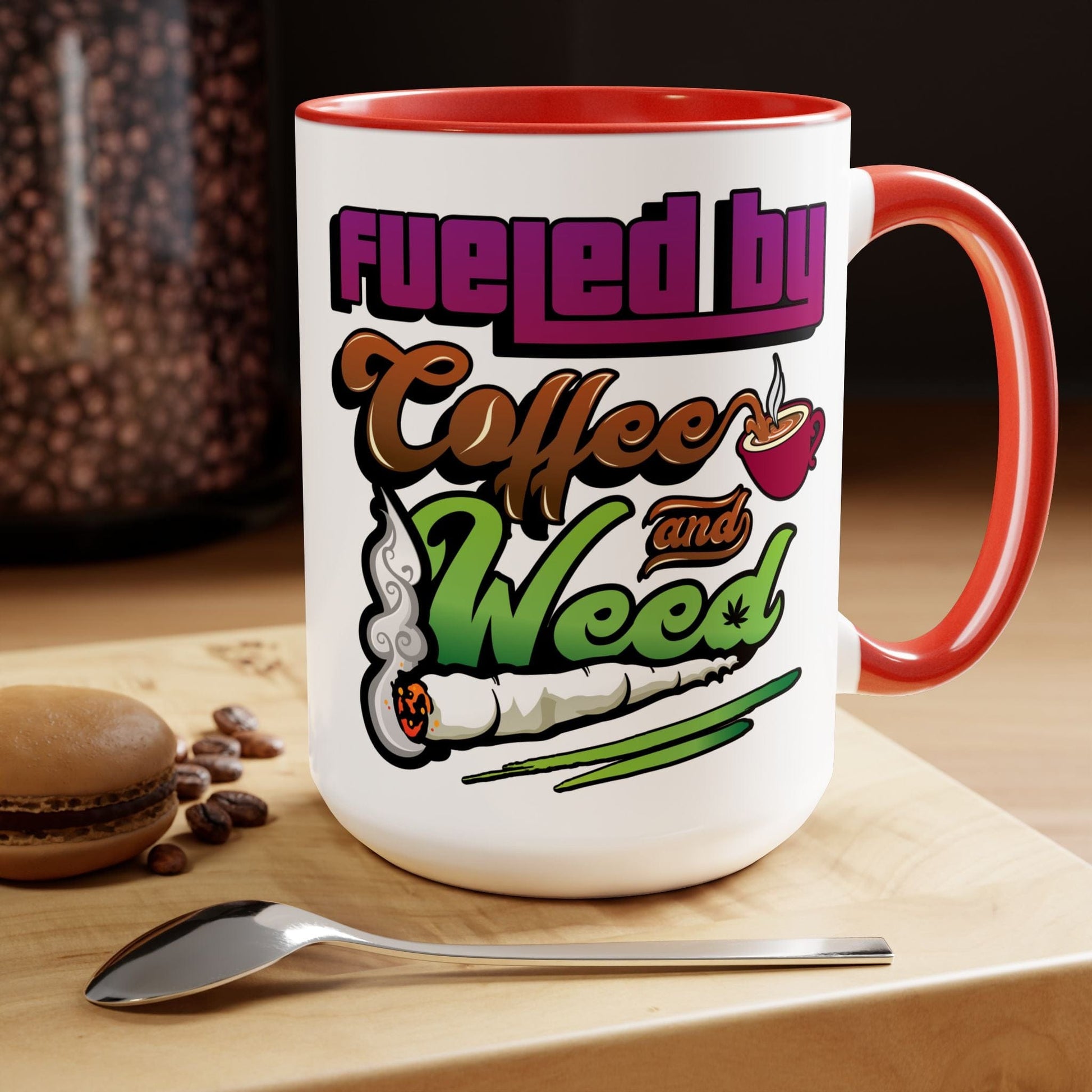 The Fueled by Coffee and Weed 15oz Coffee Mug rests on a wooden surface, accompanied by a spoon, coffee beans, and a macaron.