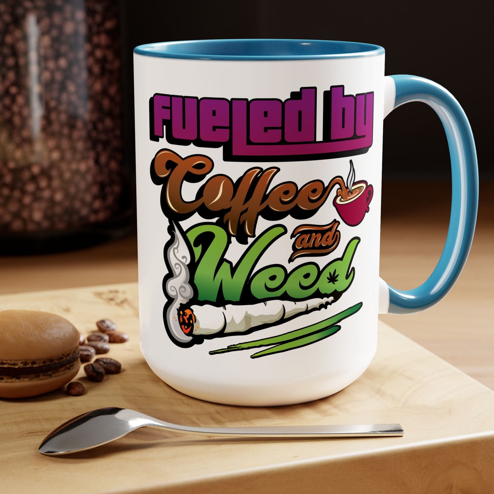 The Fueled by Coffee and Weed 15oz Coffee Mug is white ceramic with a blue handle and interior, features colorful coffee cup and joint graphics, plus the quote “Fueled by Coffee and Weed.” Pictured with a spoon, beans, and macaron.