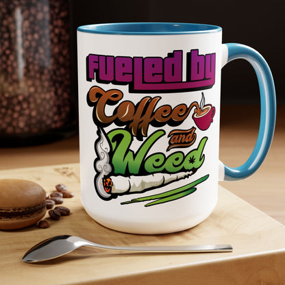 The Fueled by Coffee and Weed 15oz Coffee Mug is white ceramic with a blue handle and interior, features colorful coffee cup and joint graphics, plus the quote “Fueled by Coffee and Weed.” Pictured with a spoon, beans, and macaron.
