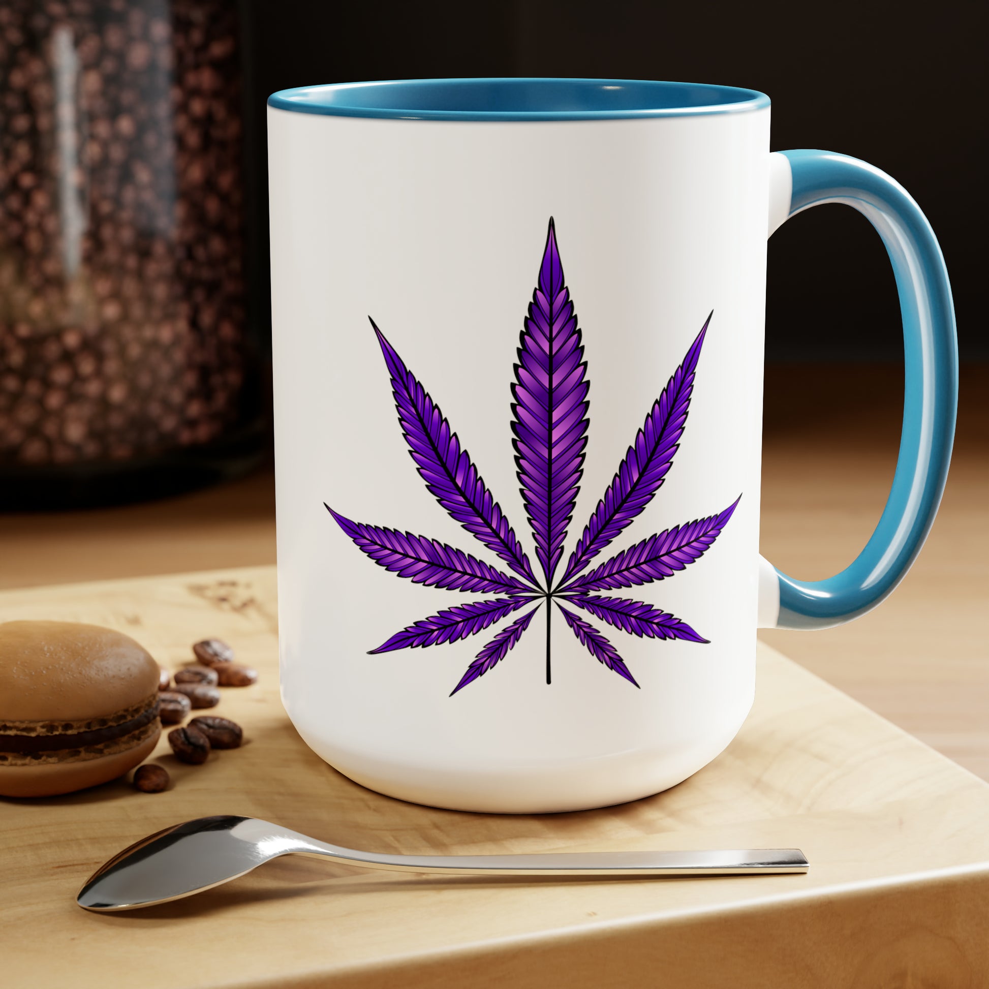 A white Purple Haze Marijuana Coffee Mug with a Purple Haze Marijuana leaf design on it, placed on a wooden table next to a macaron and coffee beans.