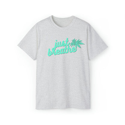 Introducing the Just Breathe Cannabis Tee: a gray t-shirt featuring the phrase "just breathe" in elegant green cursive text and a small green cannabis leaf design beside it, perfect for promoting relaxation.