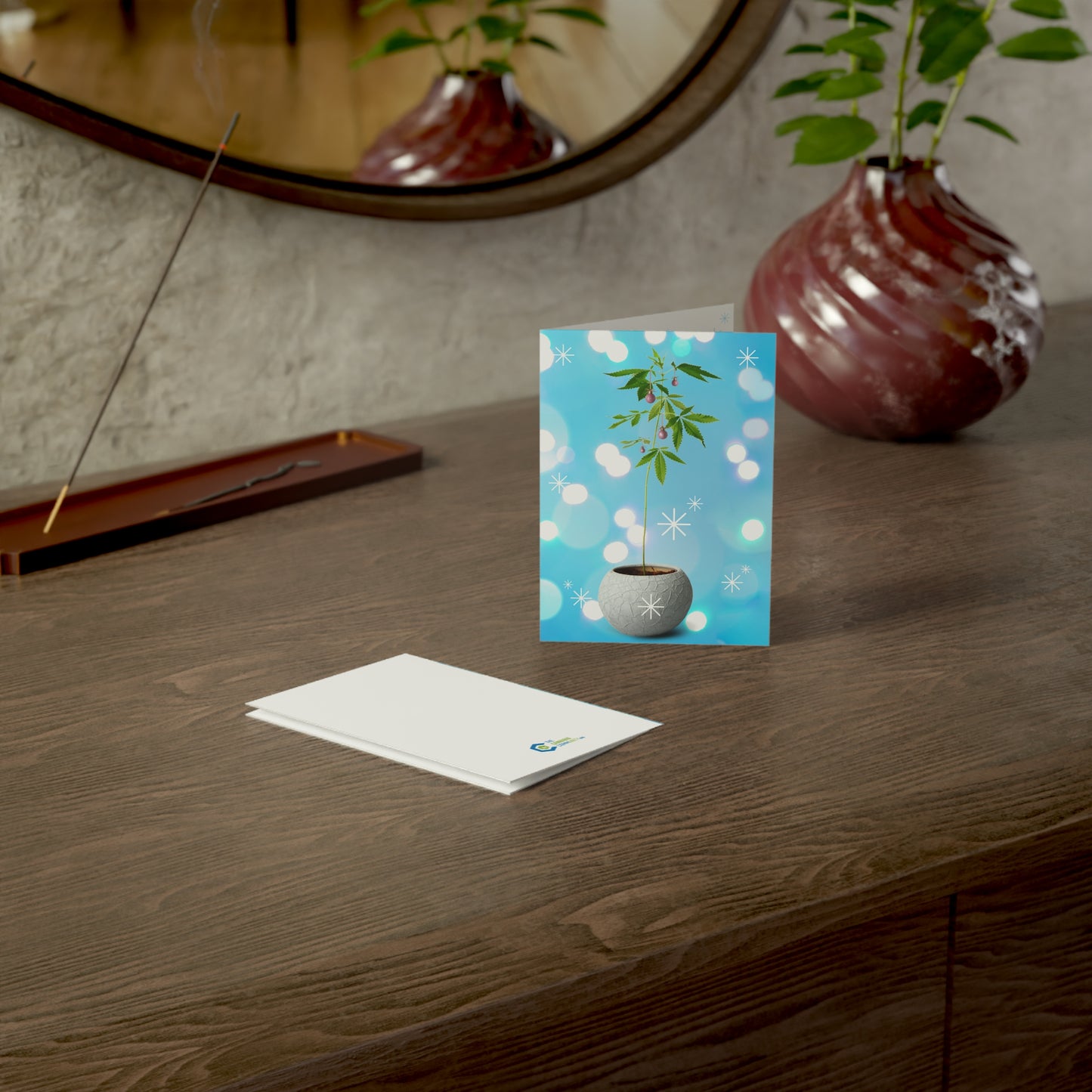 A personalized Potted Christmas Marijuana Plant Merry Christmas Greeting Card with a floral design standing on a wooden table, accompanied by an envelope, near a decorative vase with a plant.