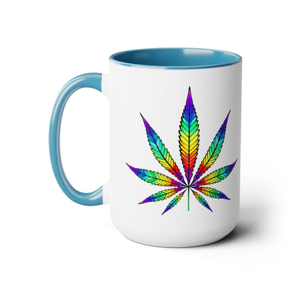 A white ceramic mug with a blue handle and interior features a colorful, rainbow-patterned cannabis leaf design on the side