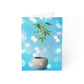 A small Christmas Marijuana plant with visible buds, growing in a spherical ceramic pot, against a bright blue background with white sparkles, ideal for featuring on sustainable paper cards.