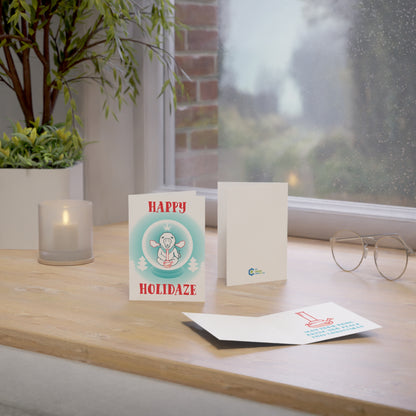 Placed on a wooden table near the window, a Happy Holidaze Zen Lion Smoking Bong Merry Christmas greeting card crafted from sustainable paper reads "Happy Holidaze," surrounded by a candle, glasses, and plant.