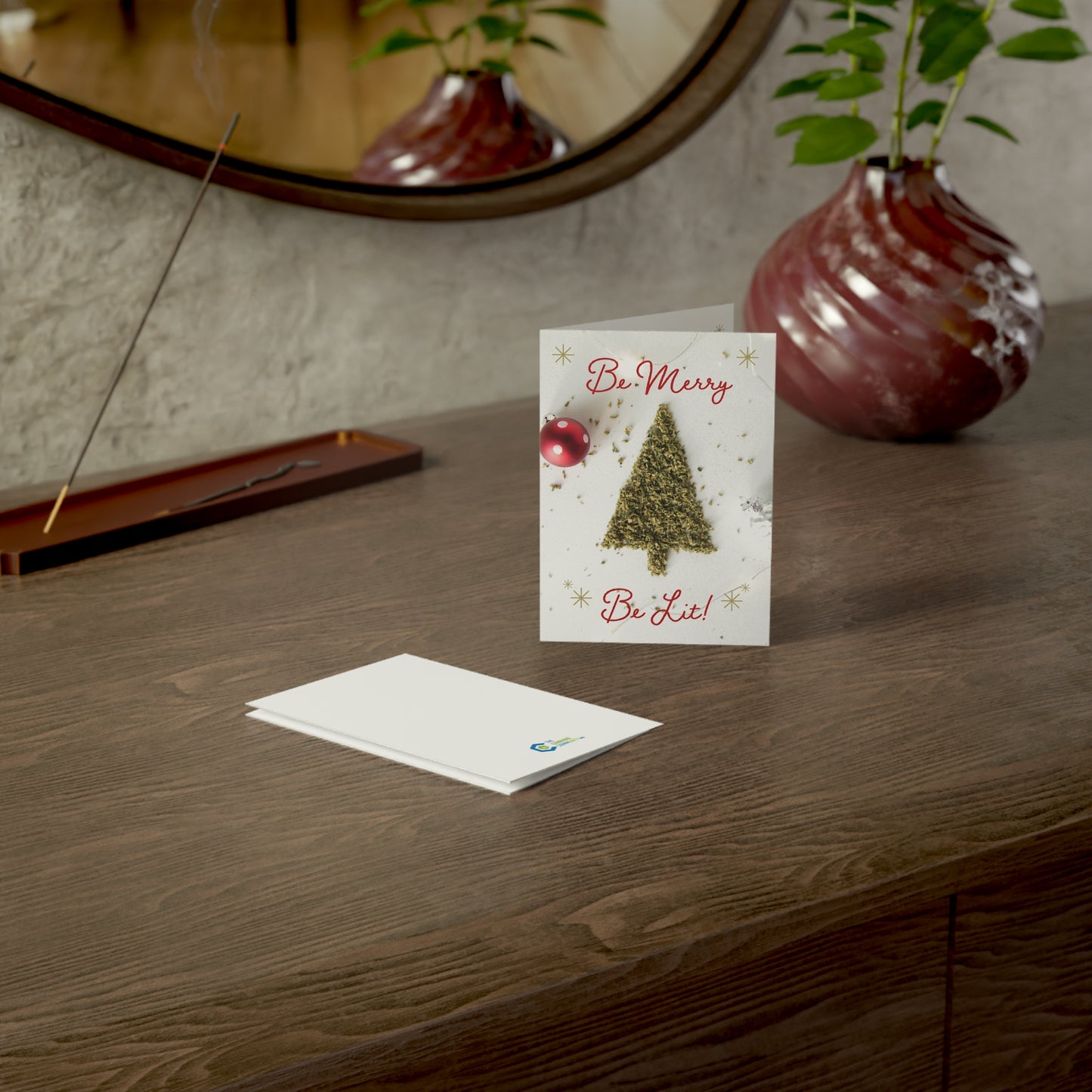 Be Merry Be Lit Christmas card being shown front, back,  laying on a table top with incense burning in the background along with a vase and mirror.