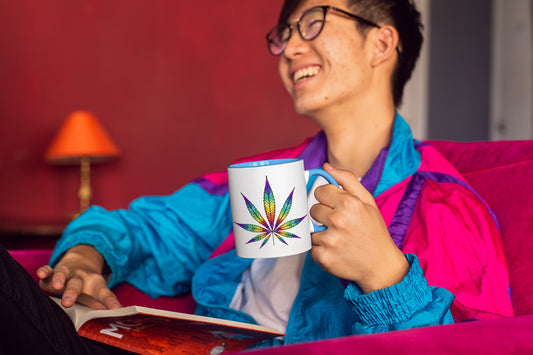 A smiling young man wearing glasses and a colorful windbreaker holds a white coffee mug featuring a vibrant, rainbow-colored cannabis leaf design. He is sitting comfortably and reading a book against a red background.