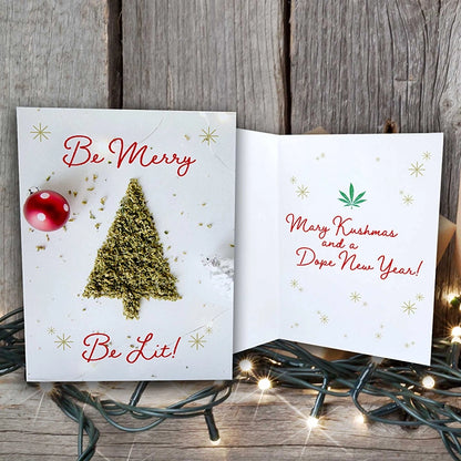 Open holiday card on rustic wood with 'Be Merry Be Lit' over a cannabis leaf tree on the front, and 'Merry Kushmas and a Dope New Year!' inside, amidst soft glowing Christmas lights.