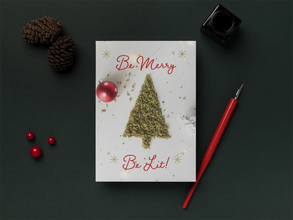 A creative Christmas card with a tree made of ground cannabis, a red bauble, and golden snowflakes, accompanied by pine cones, a red fountain pen, and an inkwell on a dark green background.