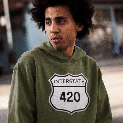 A man wearing an Interstate 420 Stoner Hoodie, featuring a green design and a white shield logo, stands outdoors, gazing to the side.