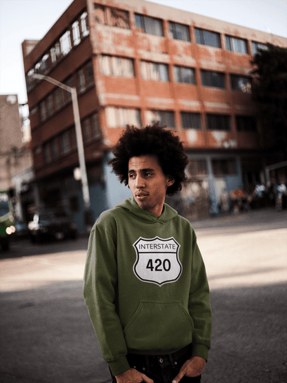 A person with curly hair wears a green "Interstate 420 Stoner" hoodie, standing on an urban street with a brick building in the background.