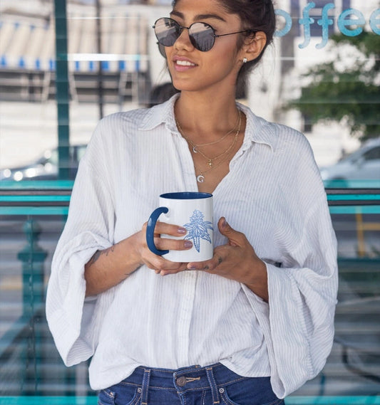 A woman wearing sunglasses, a white blouse, and blue jeans holds a Blue Dream Cannabis Coffee Mug with a blue handle and design while standing in front of a glass window.