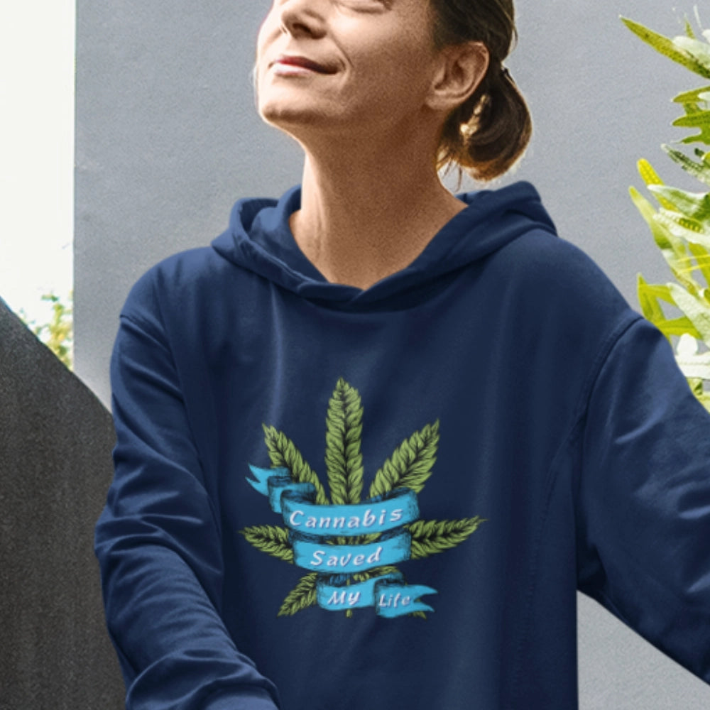 Woman in a blue Cannabis Saved My Life Marijuana Hoodie with a design saying "cannabis saved my life" surrounded by green leaves, standing outdoors, smiling with her eyes closed.