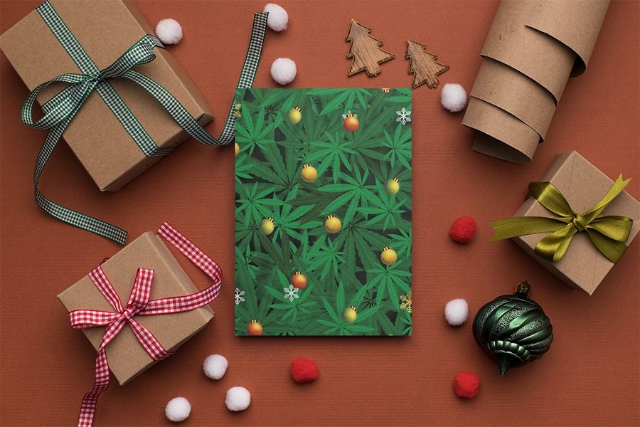 A stoner-themed Christmas greeting card mockup is displayed amidst holiday decorations. The card's design showcases a pattern of green cannabis leaves with festive ornaments and snowflakes. Surrounding the card are gifts wrapped in brown kraft paper, tied with green plaid and red checkered ribbons, and a gift with a gold bow. Small wooden tree shapes, red and white pom-poms, and a green Christmas bauble complete the holiday scene on a rust-colored surface.