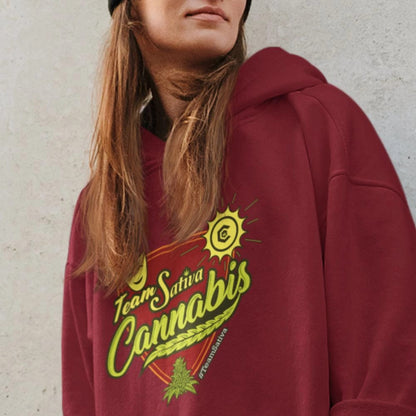 A person in a maroon Team Sativa Cannabis Stoner Hoodie stands against a gray wall.