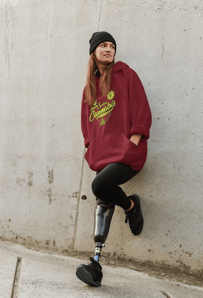 A woman with a prosthetic leg leans against a concrete wall, sporting a maroon Team Sativa Cannabis Stoner Hoodie, black pants, and a black beanie.