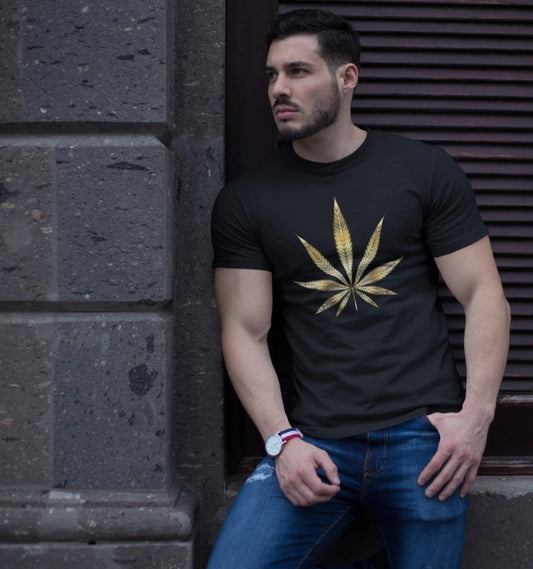 A person leans against a wall, wearing a Gold Marijuana Leaf Tee in black with blue jeans, embracing cannabis culture.