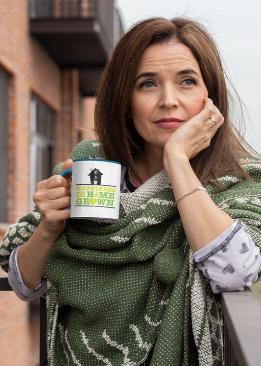 A woman sits on a balcony, holding the "Happiness Is Homegrown Weed Coffee Mug," a 15oz ceramic mug adorned with a subtle marijuana leaf design. She is wearing a green shawl and resting her head on her hand.