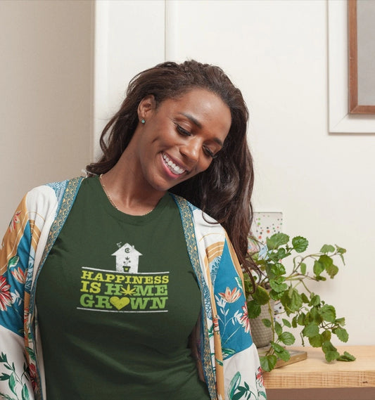 A woman wearing the "Happiness Is Homegrown Pot Shirt" in green stands indoors, smiling and looking down, with a plant in the background.