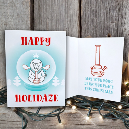 Happy Holidaze Zen Lion Smoking Bong Merry Christmas Greeting Card open on a wooden surface with a "happy holidaze" message and an illustration of a snowman in a snow globe on the left, and a festive message with a bong.