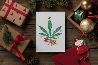 Christmas-themed image featuring a card with a Mary Kushmas + Dope New Year Marijuana Tree adorned with festive decorations on FSC certified paper, surrounded by gifts and seasonal ornaments on a wooden table.