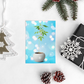 Holiday setup with a greeting card depicting a cannabis plant in a pot, set against a whimsical bokeh background, beside a chic wrapped gift, festive pine cones, and a delicate tree ornament.