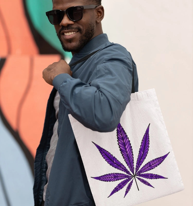 A person wearing sunglasses and a blue jacket carries the Purple Haze Marijuana Tote Bag, known for its durability and spaciousness, showcasing a white base with a striking purple cannabis leaf design.