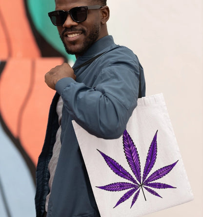 A person wearing sunglasses and a blue jacket carries the Purple Haze Marijuana Tote Bag, known for its durability and spaciousness, showcasing a white base with a striking purple cannabis leaf design.