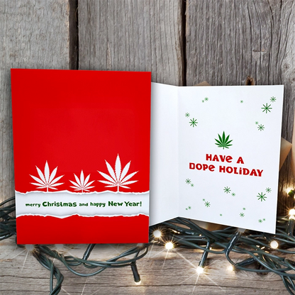 A Have a Dope Holiday Merry Christmas Happy New Year Greeting Card made of sustainable paper on a wooden table, with the cover featuring a red background and white cannabis leaves, and the inside reading "have a dope holiday" surrounded by green snowflakes