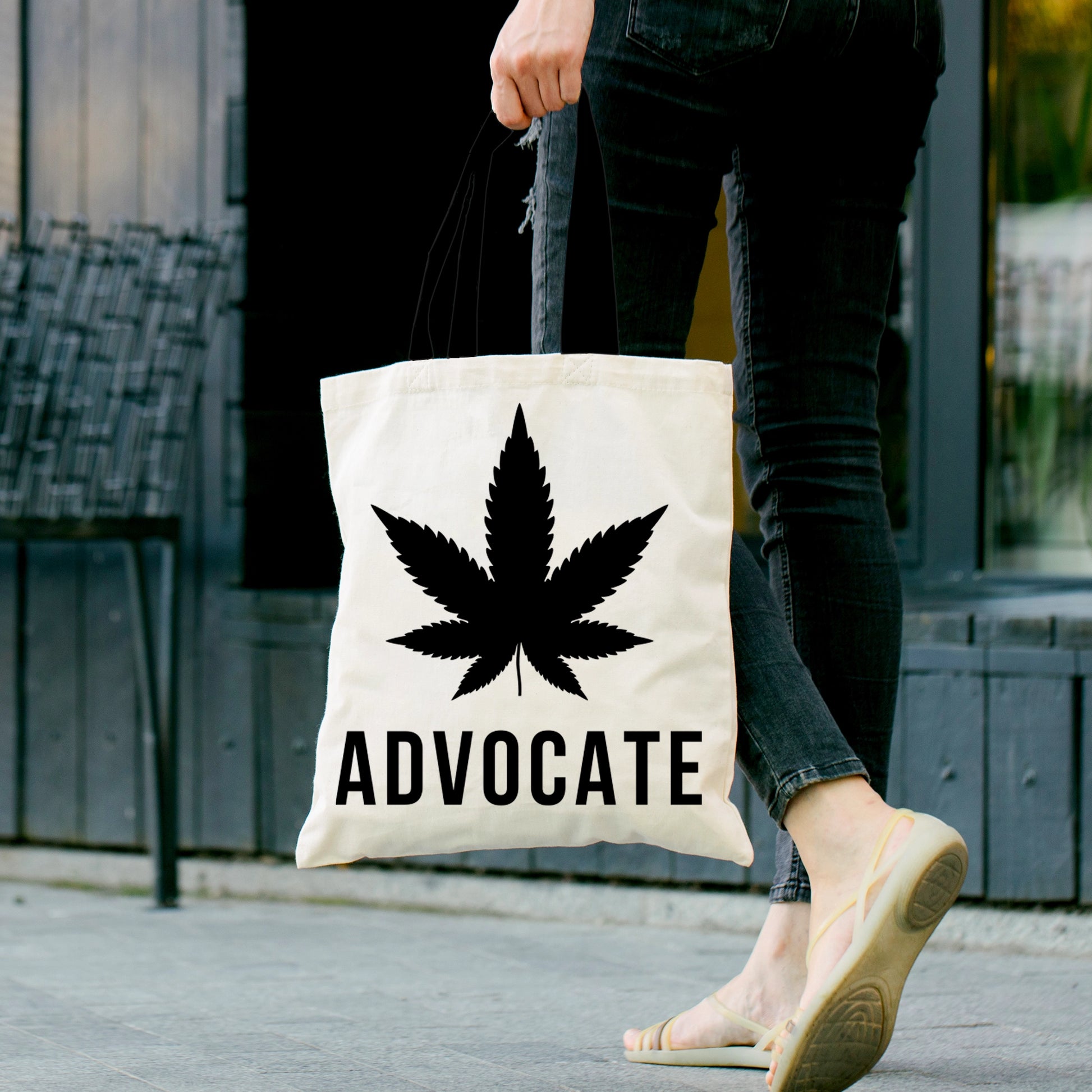 A white Cannabis Advocate Pot Leaf | Beach Grocery Tote Bag being carried by a young woman that  has a weed leaf graphic design on front and the word Advocate written in bold black lettering
