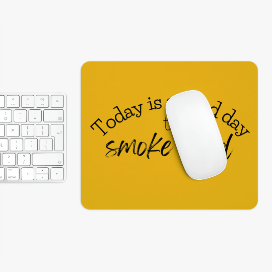 A white computer mouse on a "Today is a Good Day to Smoke Weed" yellow mouse pad, next to a white keyboard on a white surface.