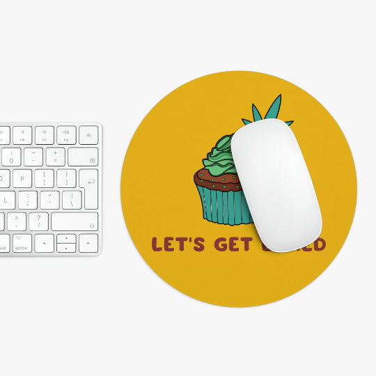 Top view of a desk with a white keyboard and a computer mouse on a Let's Get Baked Marijuana Yellow Mouse Pad featuring a graphic of a pineapple-topped cupcake with the text "let's get