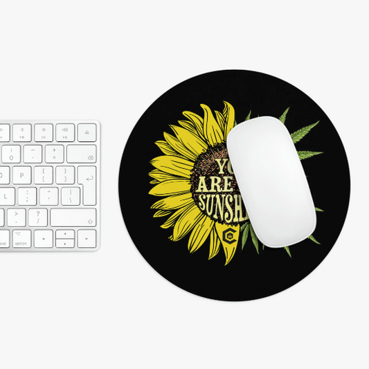 A white computer mouse rests on a round You Are My Sunshine Cannabis Black Mouse Pad featuring the text "you are my sunshine." A white keyboard is partially visible to the left.