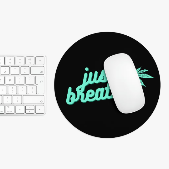 Top view of a workspace featuring a white keyboard, white mouse on a round non-slip Just Breathe Cannabis Black Mouse Pad with neon-green "just breathe" text and leaf design.