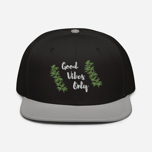 A black and gray structured Good Vibes Only Cannabis Snapback Hat flanked by green cannabis leaf designs.