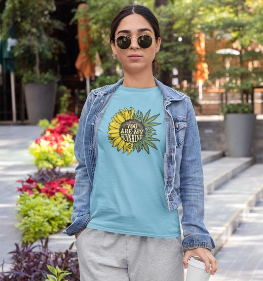 A woman stands in a garden area with potted plants and outdoor furniture, wearing sunglasses and a light blue, 100% cotton "You Are My Sunshine Cannabis Weed" shirt featuring a sunflower design. She pairs it with a denim jacket and gray pants.