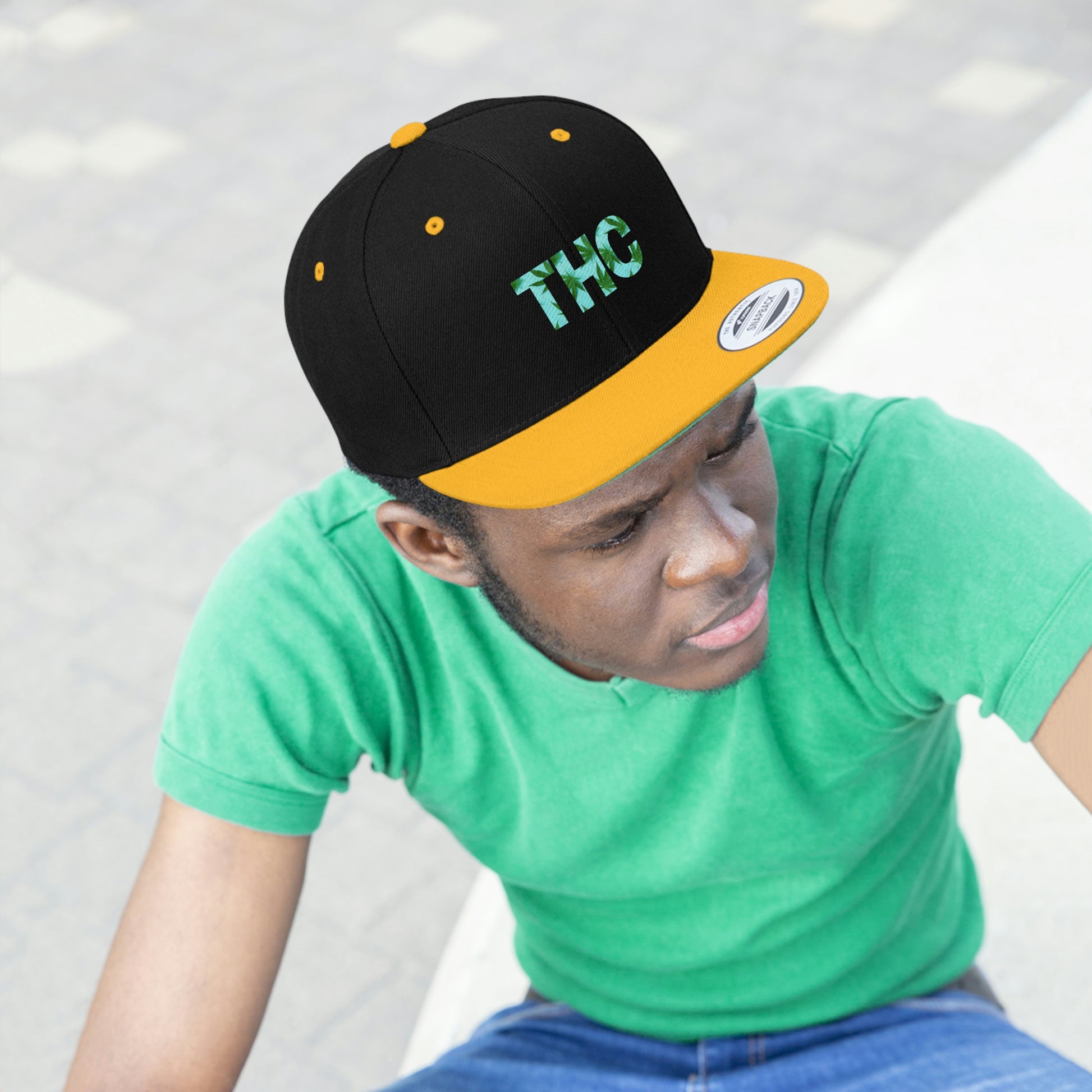 The black and gold THC Snapback Hat color way with green marijuana leaves worn by a young man in a green shirt