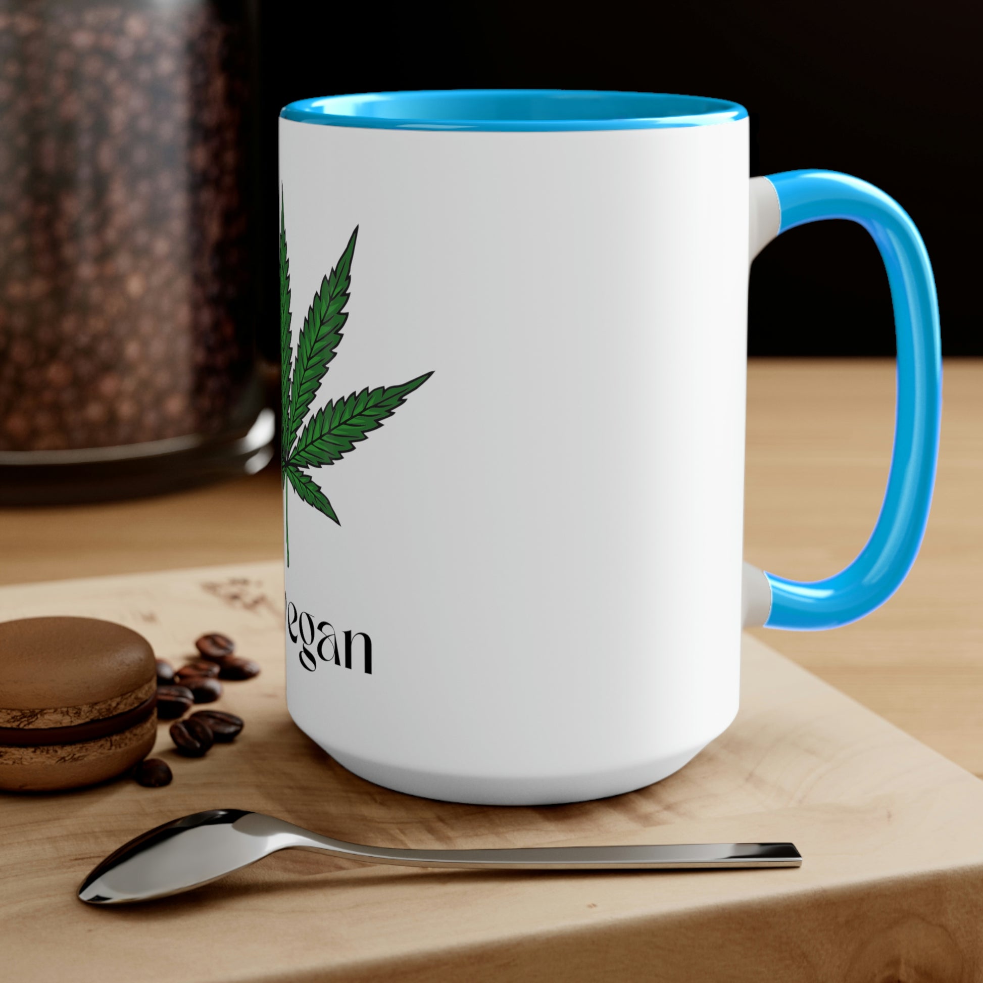 A white and light blue "I'm Vegan" cannabis coffee mug on a wooden table with a spoon and a macaroon