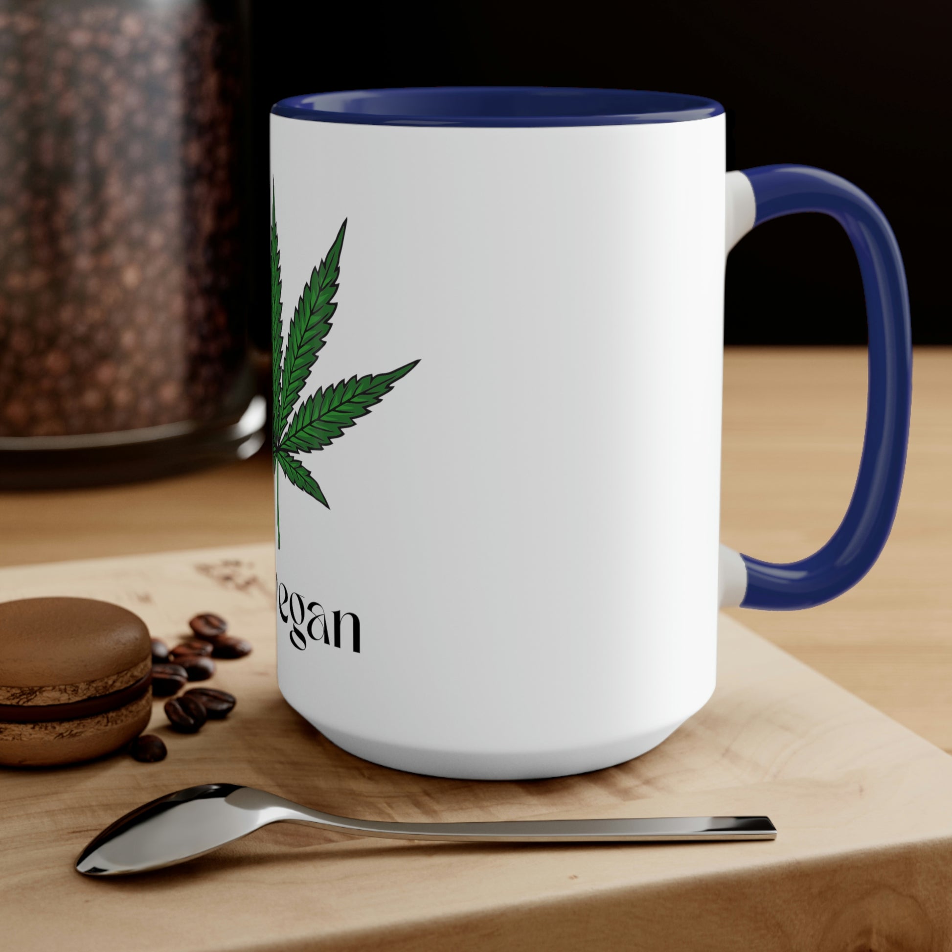 A white and navy "I'm Vegan" cannabis coffee mug on a wooden table with a spoon and a macaroon
